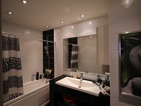 черно белая штора над белой ванной, двухцветная плитка на стенах, черная плитка на столешнице с белой раковиной у стены с большим прямоугольным зеркалом светлой ванной квартиры на первом этаже