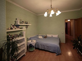 белая кровать с белыми подушками и серо зеленым покрывалом, белая напольная ваза, белый шкаф с полками в спальной комнате современной квартиры молодоженов на втором этаже многоэтажки