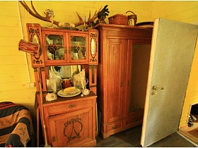 плетенные корзины на верху шкафа для одежды, оленьи рога на деревянном шкафу с посудой на полках за стеклом у стены веранды