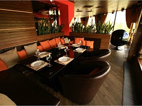 многочисленные мягкие квадратные подушки ораньжевого и бежевого цвета у спинки коричневого углового дивана и на креслах вокруг сервированного коричневого столика в уютном зале евро ресторана