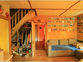 серый угловой мягкий диван с подушками в углу гостиной с открытым шкафом для одежды под деревянной лестницей стильной двухэтажной дачи