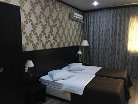 кровати с коричневым покрывалом, белыми подушками и постельным, коричневым шкафом и прикроватными тумбочками у стены с цветочными ромбовидными обоями номера гостиницы