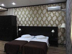 коричневый шкаф для одежды, прикроватные тумбочки у двух односпальных кроватей с широким изголовьем и белыми плафонами светильников со встроенными розетками в современном гостиничном номере