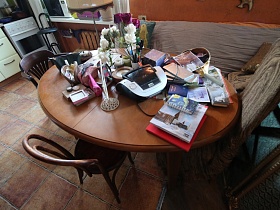 ваза с цветами,блокноты, книги, альбомы, разнообразные косметички, магнитола на поверхности коричневого раскладного стола у дивана на полу кухни с коричневой плиткой