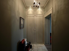 люстра, стилизованная под свечи на потолке, картина на стене и пуфик с одеждой в прихожей бежевого цвета современной дизайнерской квартиры