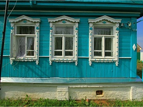 резные белые наличники на окнах старой деревянной голубой дачи СССР с белым цоколем на зеленом участке у поля