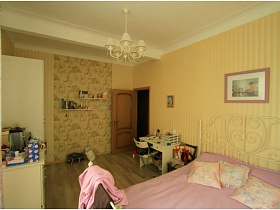 квадратные подушки на сиреневом покрывале большой кровати с металлической спинкой у стены с картиной в сиреневой рамке, деревянные полки на стене с отделочными обоями светлой гостиной во французском стиле