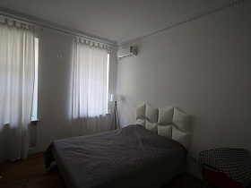 спинка из белых надувных подушек большой кровати с серым покрывалом в белой спальне с белыми гардинами на окнах скандинавской квартиры
