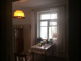 старый деревянный буфет в углу комнаты с бежевыми панелями, обеденный стол с кухонными предметами на поверхности у окна кухни с белой гардиной кв.24
