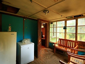 белый холодильник и стиральная машинка у стены с синим крагисом и деревянная мебель на застекленной веранде жилого домика отшельника среди новостроек