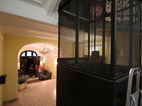 лестница-стремянка у лифтовой кабины за черной сеткой на первом этаже парадного подъезда с жилыми квартирми, диваанами у желтых стен коридора с арочными входными дверьми
