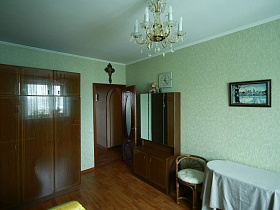 зимний пейзаж на картине над овальным столом, квадратные белые часы, церковный крест над входной дврью в бирюзовуй комнату яркой современной трехкомнатной квартиры с комнатой бабушки