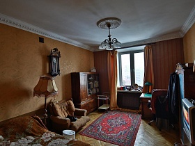 старинные часы с маятником над бежевым мягким креслом, торшер с бежевым абажуром,книжный шкаф и письменный стол у окна с персиковыми шторами в спальне советской трехкомнатной квартиры