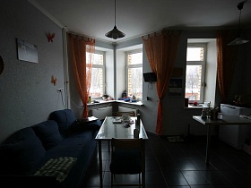 ораньжевые гардины на эркерных окнах кухни с серыми стенами, синим мягким диваном у обеденного стеклянного стола трехкомнатной современной квартиры