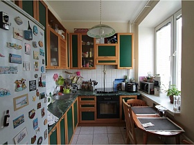 мраморная зеленая плита в коричневой кухне с зелеными дверцами,раздвижной обеденный стол у окна и белый с зеленым плафон люстры на потолке светлого помещения кухни в простой квартире на Садовом