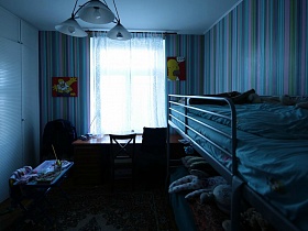 большой письменный стол у окна с голубой гардиной в детской спальной комнате с полосатыми обоями в семейной трешке