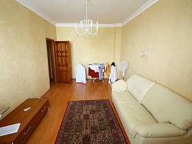 белый мягкий диван с подушками, цветной красно-коричневый коврик рядом и односекционный комод на ножках светлой гостиной трехкомнатной квартиры в переезде (въезде) молодоженов