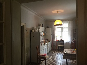 желтый абажур подвесной люстры на белом потолке светлой кухни с белой мебельной стенкой, холодильником, деревянными стульями и обеденным столом на полу с шахматной плиткой