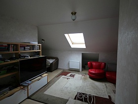 небольшая мебельная стенка, красное круглое мягкое кресло на светлом цветном ковре в комнате отдыха в мансарде жилого дома