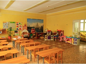 учебные пособия на стене, полочках и игровая зона просторной комнаты в детском саду