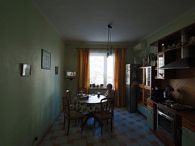 стулья со спинкой вокруг круглого обеденного стола с цветной клеенкой напротив окна с белой гардиной и  желтыми шторами на кухне большой семейной квартиры в Марьино