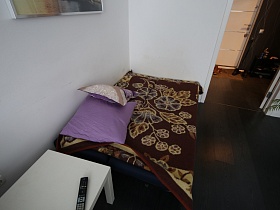 пульт от телевизора на белом столике у тахты с сиреневыми подушками на цветном коричневом покрывале в светлой гостиной с открытой дверью в прихожую современной просторной квартиры