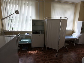 холодильник, медицинский столик, белый шкафчик для медикаментов, кушетка за ширмой у окон с жалюзями в медицинском кабинете спортивной школы
