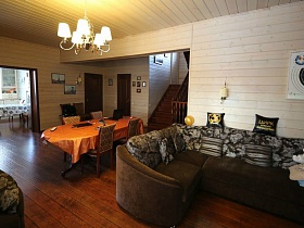 большой коричневый угловой диван с цветными подушками и стол с ораньжевой скатертью по обе стороны открытого дверного перехода на деревянную лестницу современного дома в три этажа