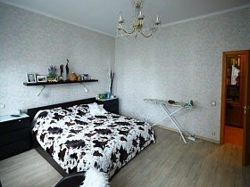 настенная полка с фотографиями, статуэтками над большой кроватью в спальне с белыми стенами трехкомнатной современной квартиры