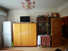 белый холодильник, желтый трехдверный шкаф с телевизором наверху, сушилка для белья у большого книжного шкафа с куклами и мягкими игружками сверху у стены спальной комнаты с кондиционгером сталинской квартиры