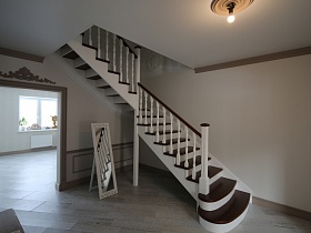 белый потолок и кремовые стены просторного вестибюля с напольным зеркалом под красивой двухцветной деревянной лестницей на второй этаж современного коттеджа