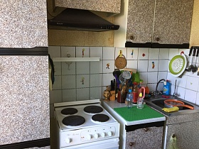 коричневая вытяжка над белой газовой плитой между шкафами светлой мебельной стенки с дверцами под мрамор, кухонным инвентарем на шкафу у мойки кухни трехкомнатной квартиры