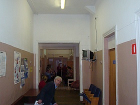 телевизор на стене длинного коридора с деревянными креслами у кабинетов, плакатами над письменном столом в здании деревянной больницы