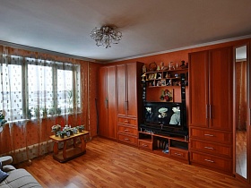 яркая рыжая мебельная стенка с многочисленными шкафами, ящиками,открытыми полками и угловым шкафом для одежды с зеркальными дверцами в гостиной с ораньжевыми стенами и белым потолком трехкомнатной квартиры с комнатой бабушки