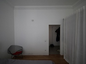необычное сетчатое кресло с красной подушкой в углу светлой спальни с открытым дверным проемом в прихожую скандинавской квартиры