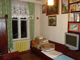 белая короткая гардина и зеленые шторы на окне,швейная машинка в футляре и многочисленные коробки на вишневом диванчике, телевизор на тумбочке у белой стены с картинами светлой комнаты бабушкиной квартиры