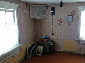 рамки с фотографиями, телевизор на деревянном полу комнаты с иконами в парадном углу, электросчетчиком на стене с цветными старыми обоями