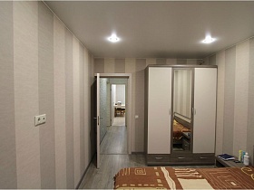 серый шкаф с белыми дверцами и зеркалом по центру у стены спальной комнаты с обоями в широкую серую полоску в семейной новостройке