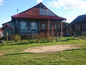 двухцветный деревянный дом на два хозяина под треугольной крышей с резными наличниками на окнах за забором из металлической сетки