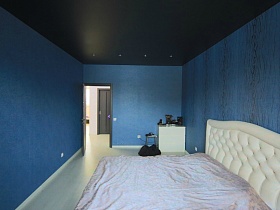 большая белая кровать с кремовым покрывалом у стены синей спальной комнаты с черным потолком молодежной двухкомнатной квартиры