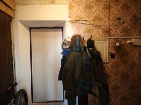 велосипед у коричневого шкафа, одежда на крючках настенной вешалки у входной двери в прихожую со старыми, поврежденными обоями советской эпохи