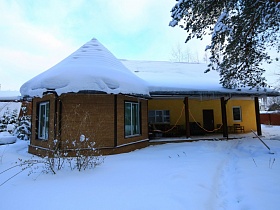 общий вид художественной дачи с шапками снега на крыше на заснеженном участке за высоким забором в сосновом лесу