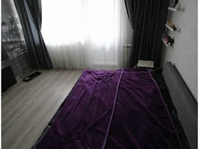 серый мягкий разложенный диван с фиолетовым покрывалом у стены с белыми деревянными полками, белой прозрачной гардиной и темно серыми шторами на большом окне с балконой дверью гостиной современной однокомнатной квартиры