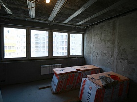 блоки строительного материала на полу освещенной комнаты с бетонными стенами и большим окном просторной двухэтажной квартиры без ремонта для съемок кино