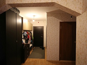большой коврик у входной двери, настенная вешалка в коричнево-бежевой мебели прихожей с большим арочным переходом  трехкомнатной квартиры после переезда