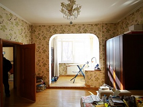 цветочная люстра на белом потолке, большой темно-коричневый шкаф для одежды в углу спальной комнаты, совмещенной с застекленной лоджией простой семейной четырехкомнатной квартиры