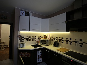 встроенная газовая плита, раковина, тостер, микроволновка, чайник и деревянная доска на светлой столешнице двухцветной кухни с плиткой на рабочей стене