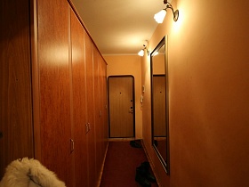 обувь на коричневой дорожке до входных дверей в прихожую с коричневым шкафом, зеркалом и бра на персиковой стене двухкомнатной квартиры