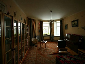 книжные шкафы со стеклянными дверцами у стены гостиной с темными шторами на эркерных окнах современной трехкомнатной квартиры