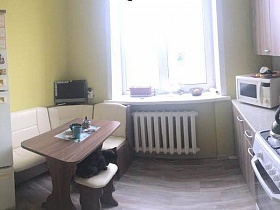 двухцветный кухонный гарнитур с коричневым обеденным столом, табуретками, телевизором на полке светлого углового дивана, газовой плитой между шкафами на кухне квартиры СССР